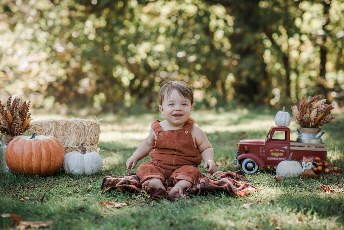 1 year old baby boy toddler in front of fall cake smash setup with hay pumpkins, little red truck, and wearing burnt orange overalls outside