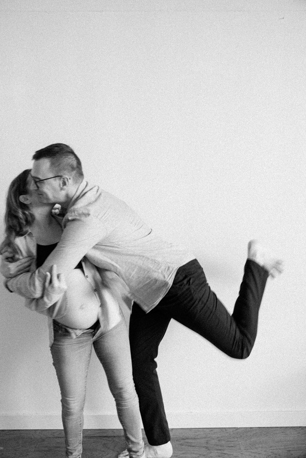 Husband playfully hugs his pregnant wife while kicking his leg in the air joyfully. 