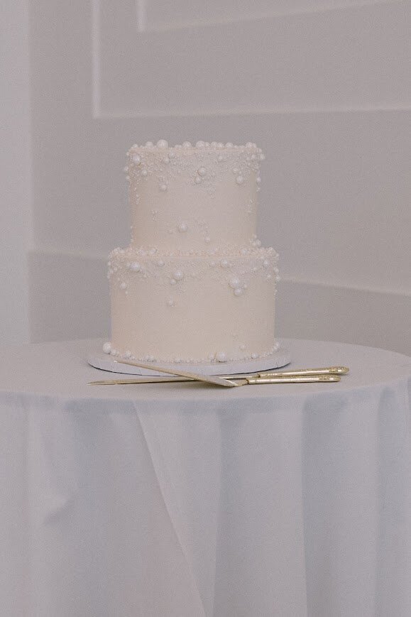 Photo shows a shot of a cocktail table with a white tablecloth, and a two tier wedding cutting cake on top. The cake is all white, with sugar pearls edging the top and bottom of each tier.