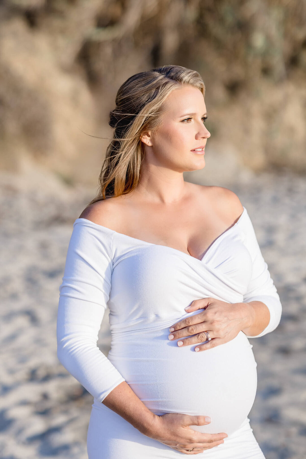 A pregnant woman in a white off-the-shoulder dress stands on a sandy beach, gazing into the distance. She gently cradles her belly with one hand above and the other below. The background is blurred with natural tones.