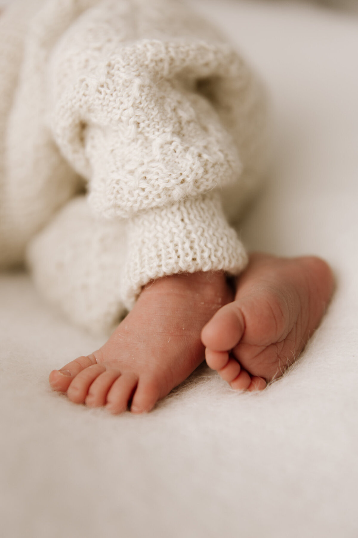A close-up of tiny newborn feet peeking out from soft, cream-coloured knit pants. The image captures the delicate wrinkles and peach fuzz of fresh baby skin, styled in a cozy and minimal studio setting.