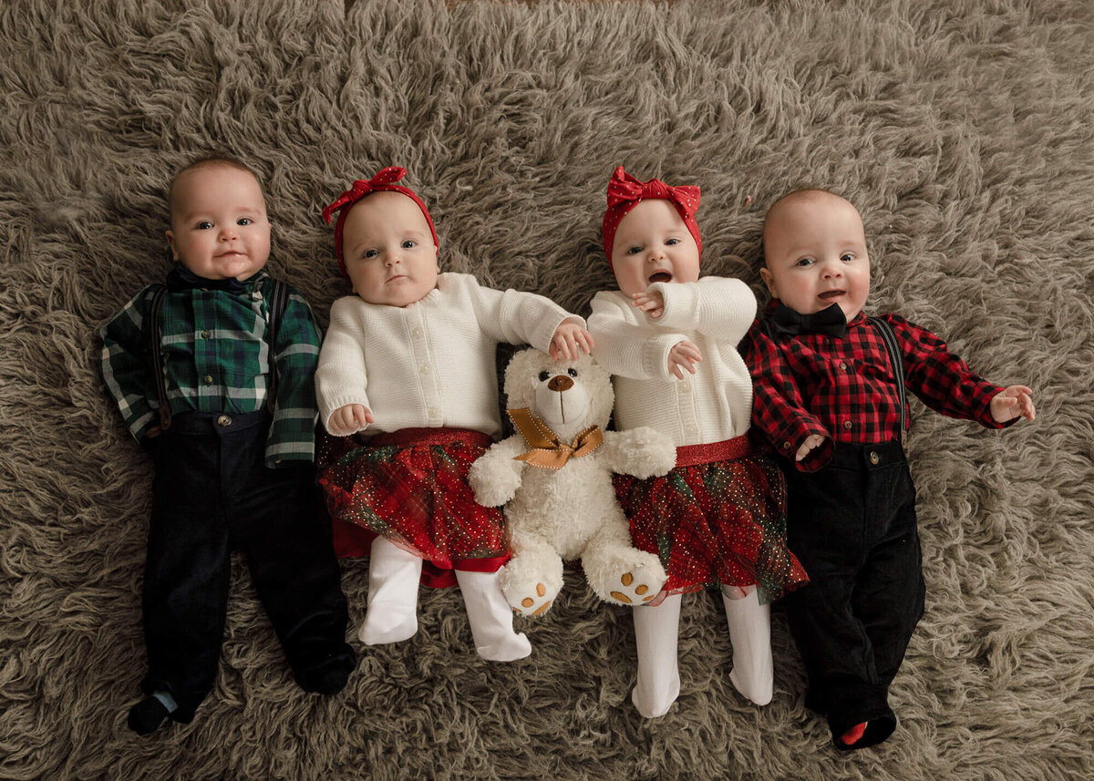 Quadruplets at 6 months old dressed in Christmas outfits laying on a gray rug with a stuffed bear included.