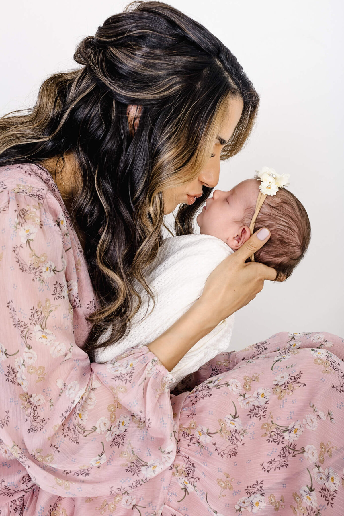 A woman in a floral pink dress gently kisses the forehead of a baby wrapped in a white blanket. The baby wears a floral headband. They are against a plain white background.
