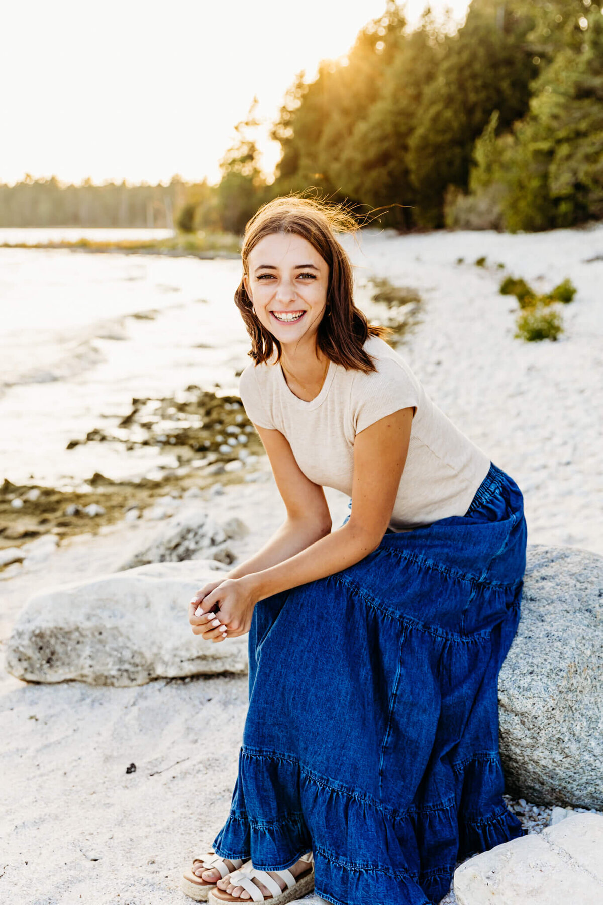 pretty teen girl in a cream top and denim skirt sitting on a large rock as she leans forward and laughs captured by senior photographer Ashley Kalbus