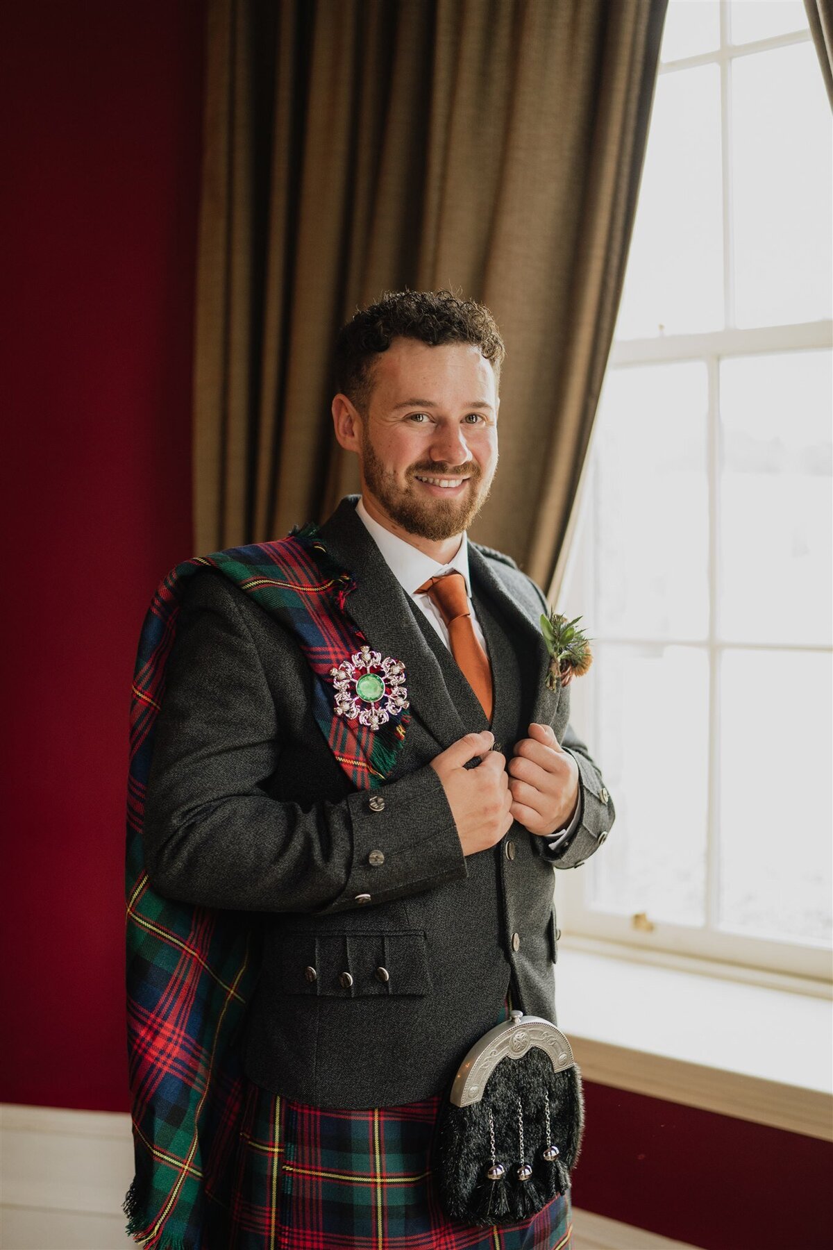 A groom stands by a large old window and smiles candidly as he gets his photograph taken. The courtyard of Logie Country House is visible through the window. The groom wears traditional Scottish attire.
