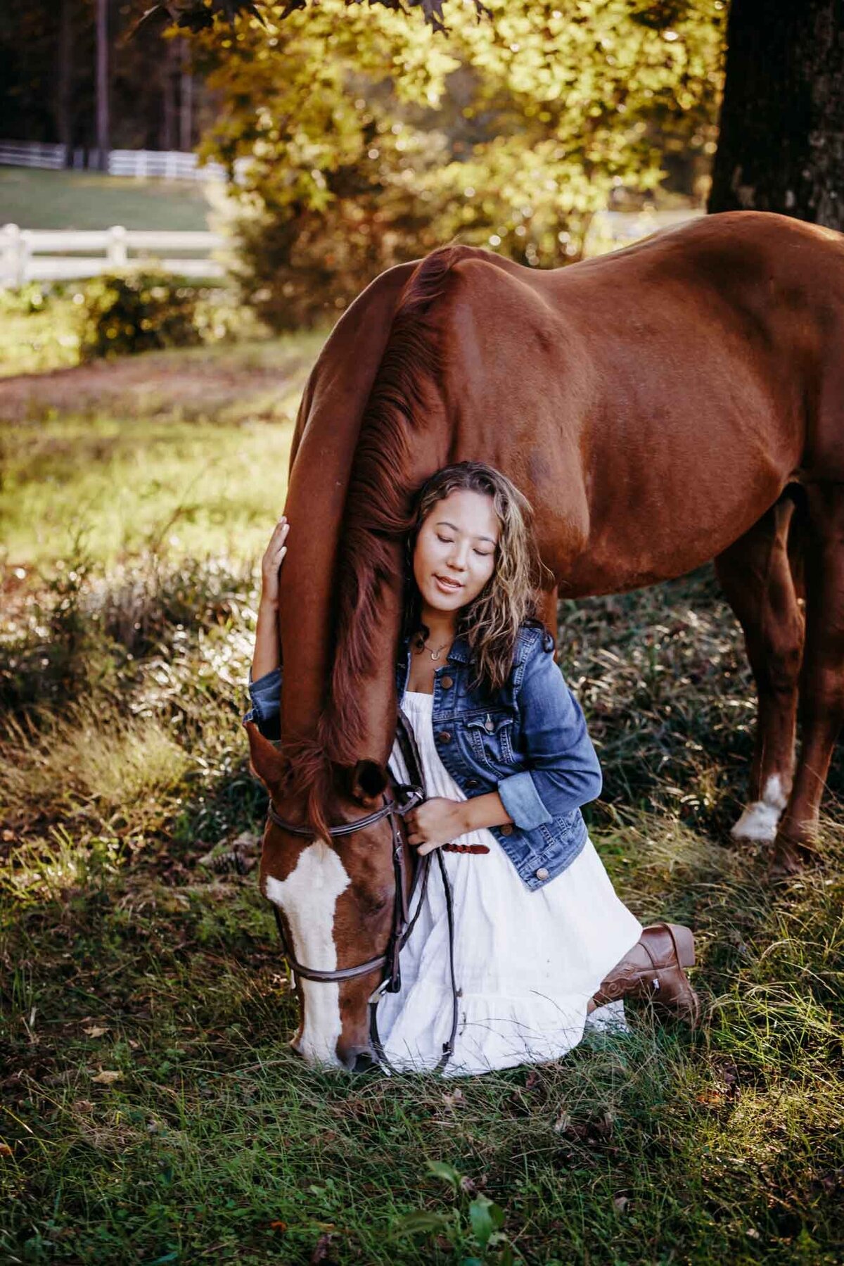 Senior portrait of a girl in a white dress kneeling on the grown next to her horse while he eats grass.