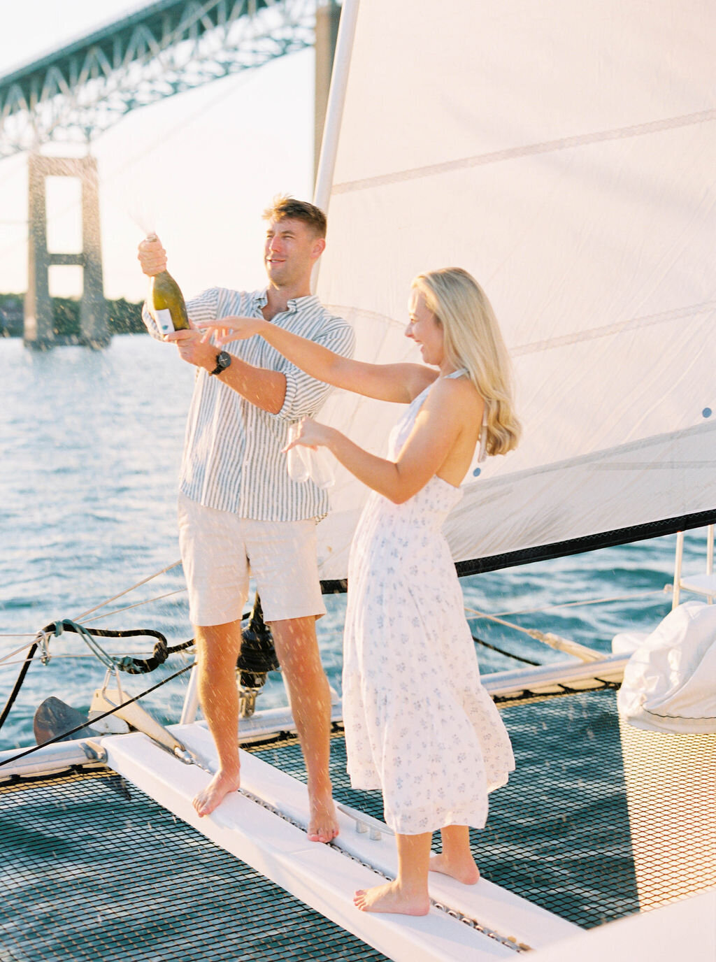 Newport Rhode Island Engagement Photo Photographer | A couple joyfully pops champagne on a sailboat, with a sunlit sea and a bridge in the background. They appear happy and celebratory, dressed in light summer clothes.