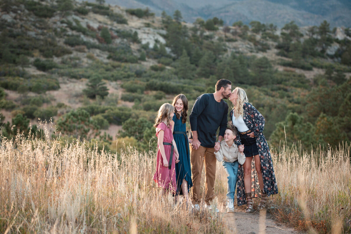 Happy parents kiss while walking up a mountain trail with their three kids for a Denver family photographer