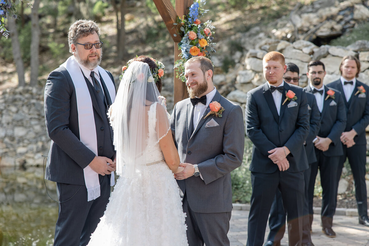 groom smiles at bride at Hayes Hollow at Hidden Falls in Spring Branch Texas under orange roses alter