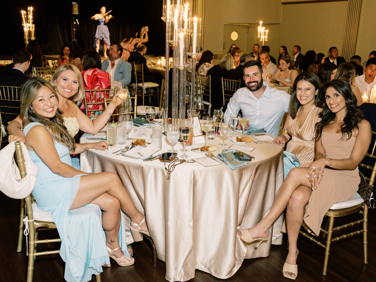 This lively image captures a group of friends sitting around a beautifully set dinner table, enjoying food and drinks, surrounded by warmth and celebration under soft lighting.