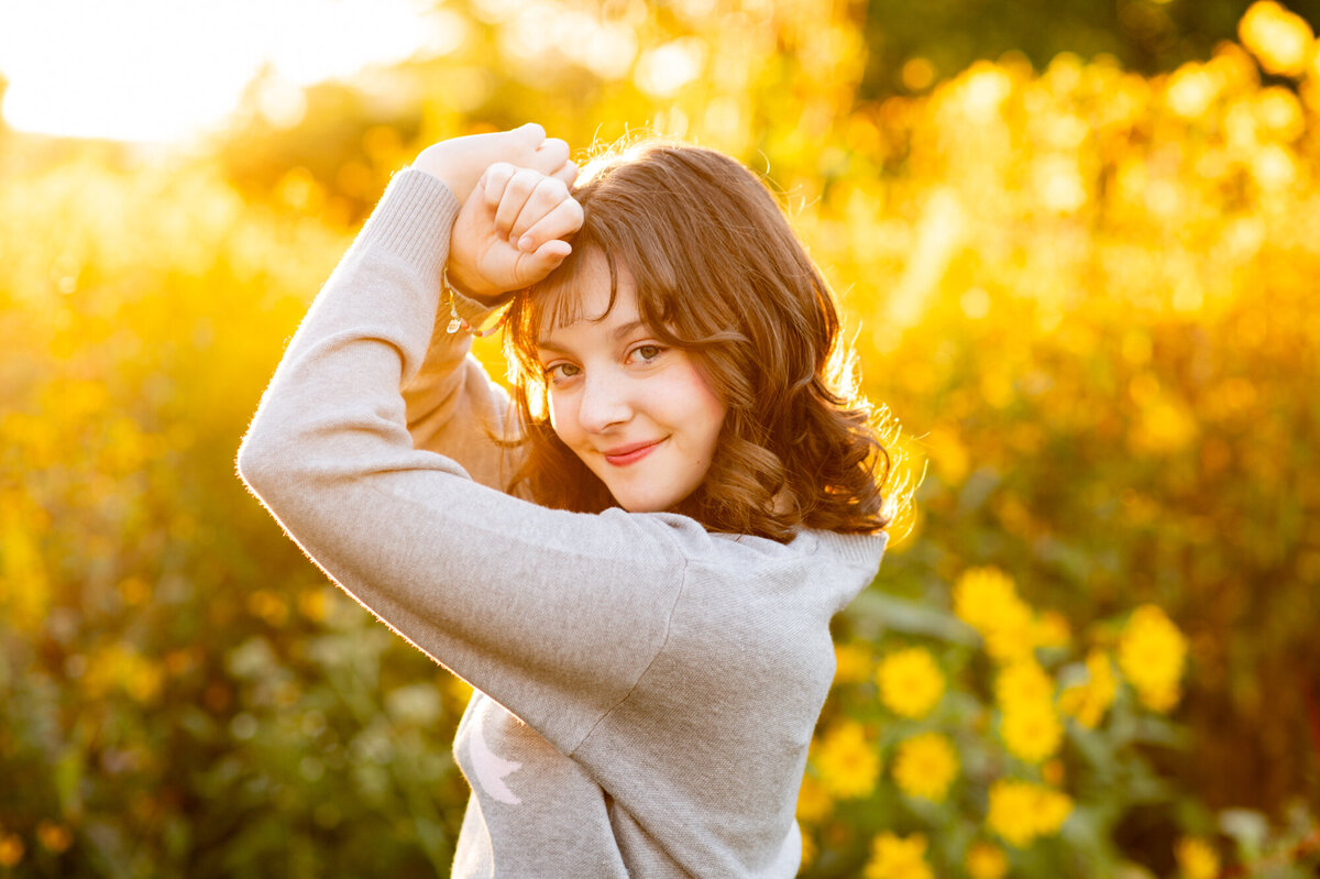 Golden hour portrait of a high school senior girl standing near a wall of yellow flowers. 