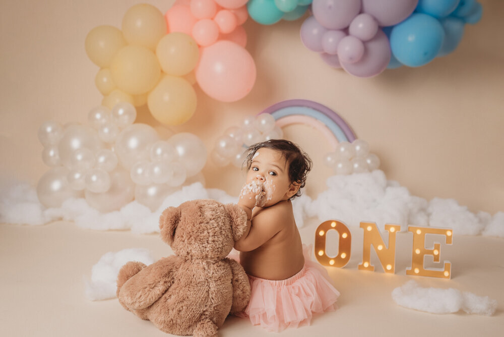 photo shoot with Atlanta, Georgia cake smash photographer with one year old baby girl sitting on tan backdrop wearing pink tutu eating cake and looking over shoulder with teddy bear sitting next to her. Rainbow pastel balloons in background.