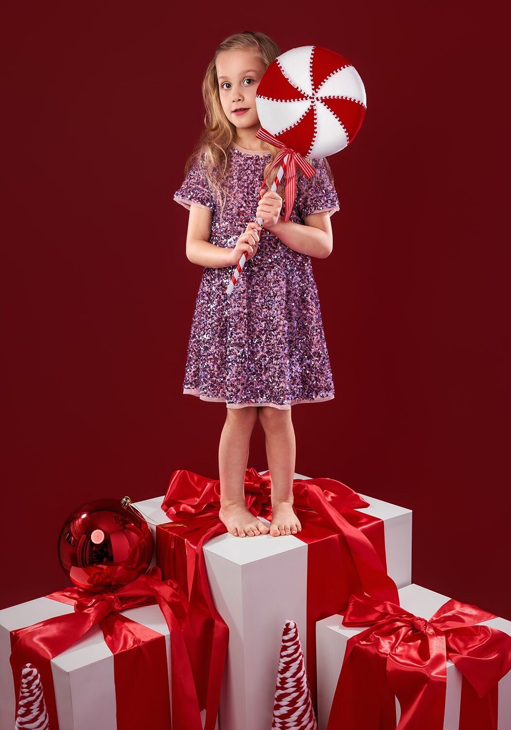 Image of a girl standing on a large present on a red Christmas backdrop holding a large lollipop in the studio. Taken by Norwich portrait photographer Claire Howes. Christmas Mini Sessions Norwich.