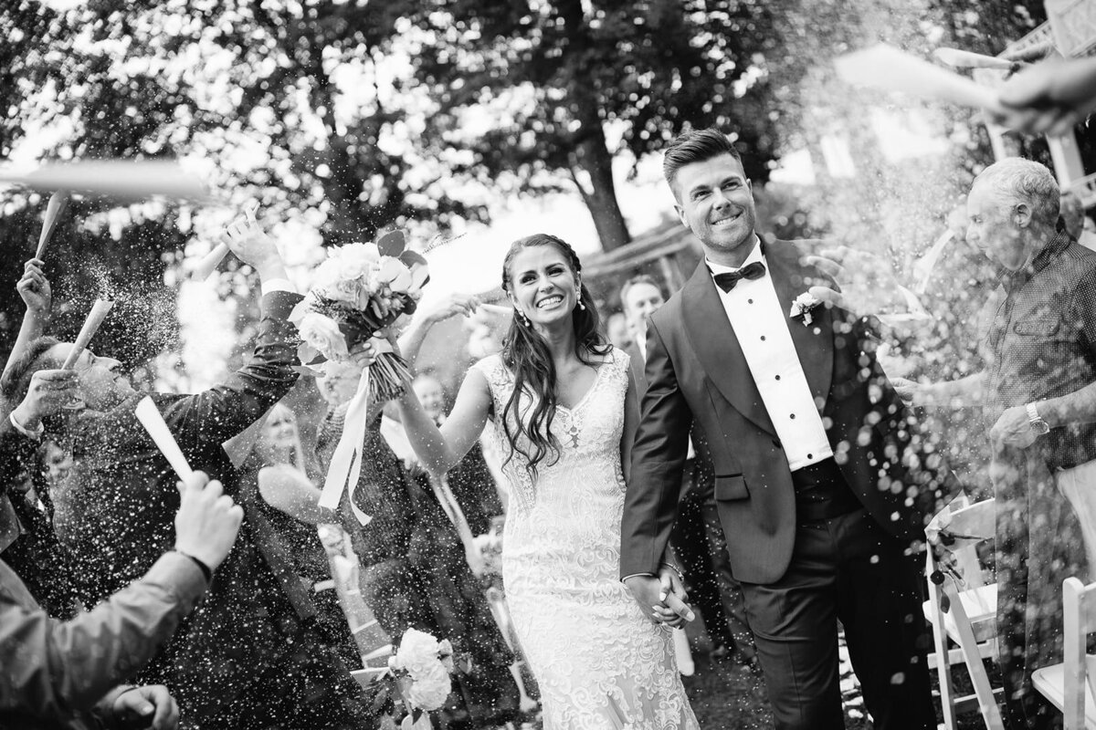 Couple de mariés sous les confettis lors de la cérémonie de leur mariage extérieur au Mnaoir Hovey, en Estrie, photo en noir et blanc. 