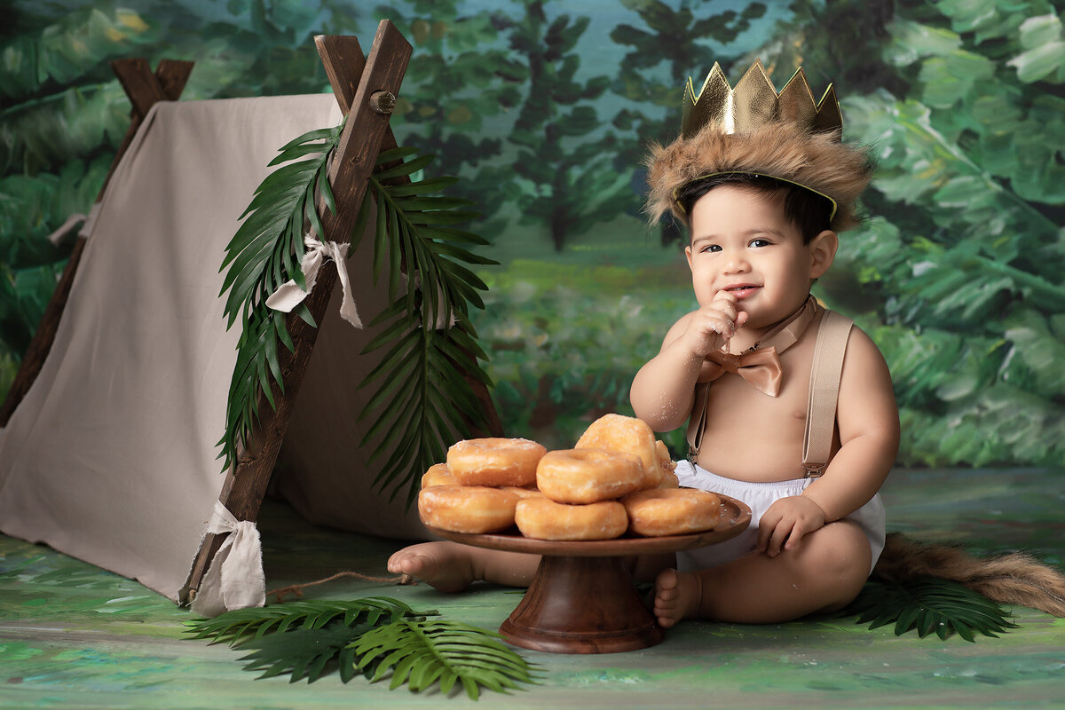 A joyful baby wearing a fur-trimmed crown and bow tie sits next to a rustic tent decorated with greenery, enjoying a pile of glazed donuts in a whimsical, forest-themed setup. Captured by a Dallas baby milestone photographer, this adorable scene embodies the playful and adventurous spirit of a one-year milestone session.