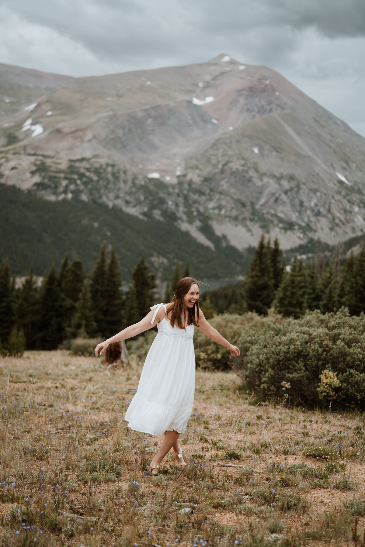 Woman twirling in white dress in front of Hoosier Pass mountain range during summer engagement session.