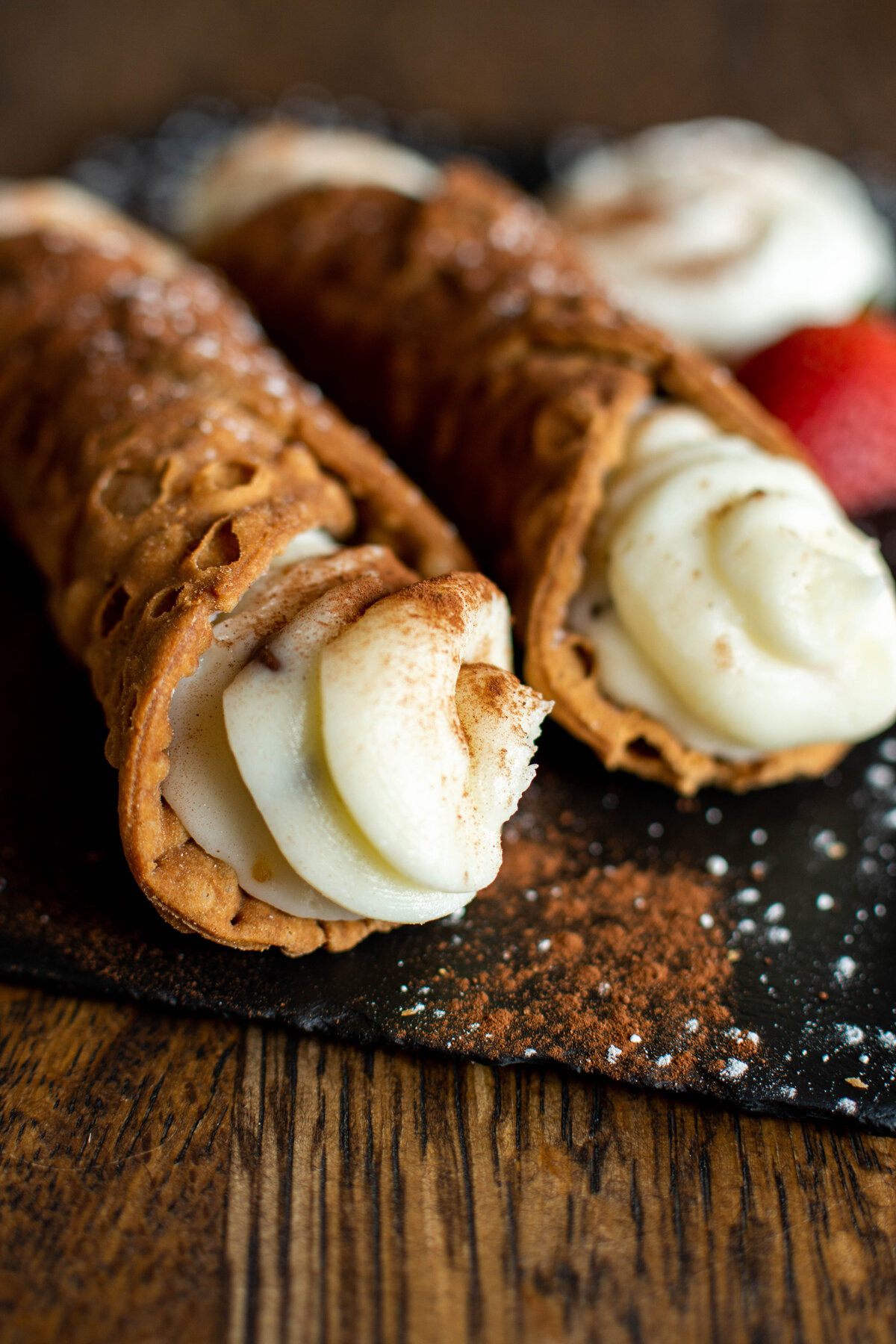 Close up image of two cannoli on a black plate with whip cream and strawberry in the background