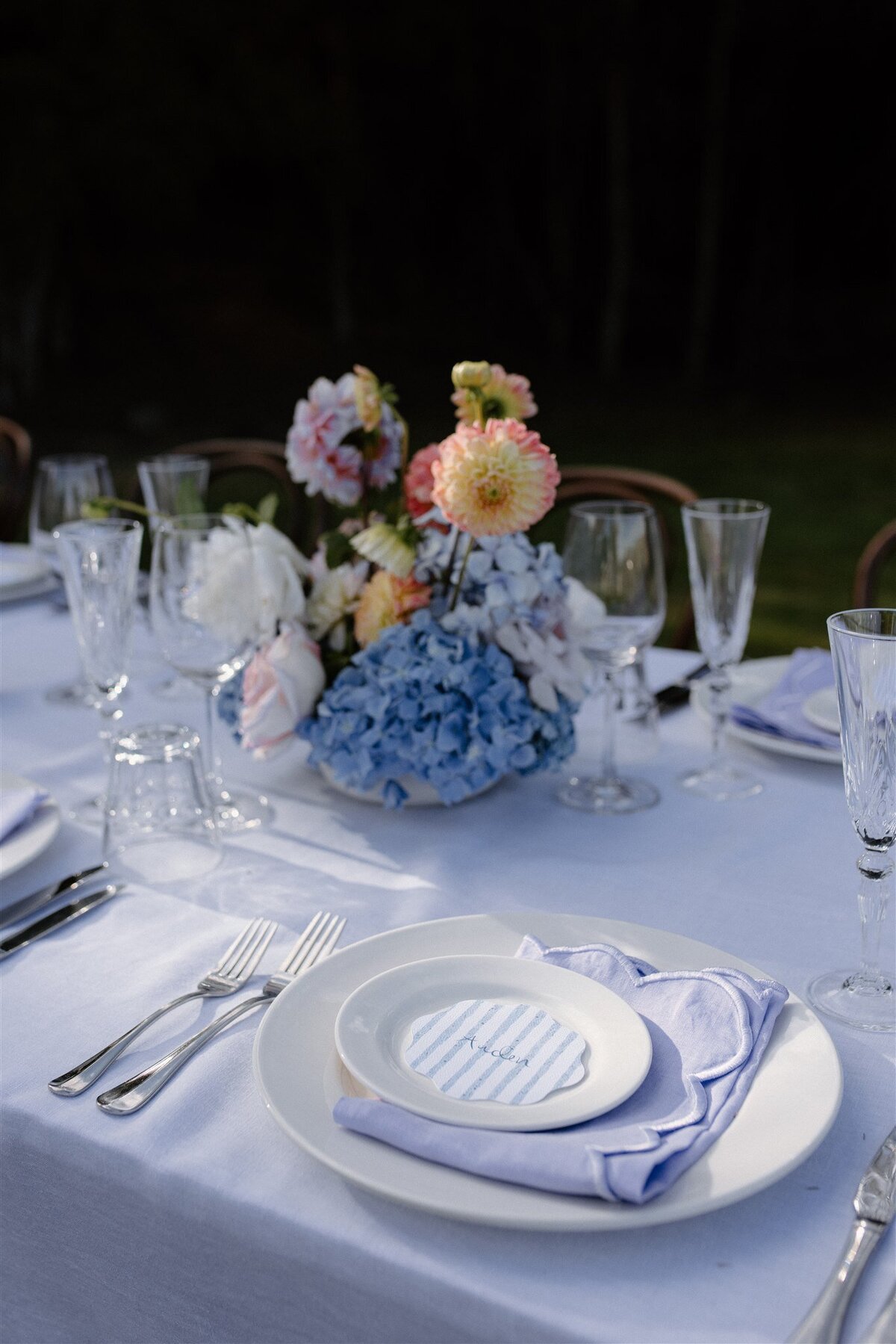 Place setting featuring blue striped name card, lilac napkin, and layered white plates on an elegant alfresco wedding table in Central Otago.