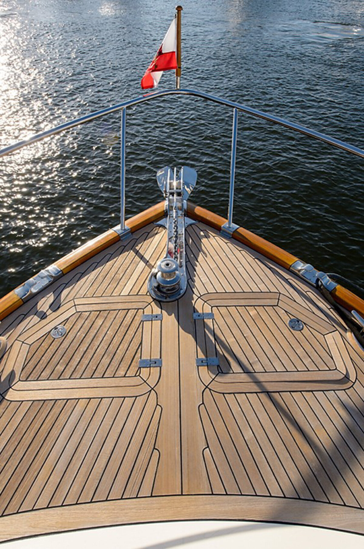View from the bow of a yacht moving through the water, showcasing a wooden deck and a French flag fluttering in the wind