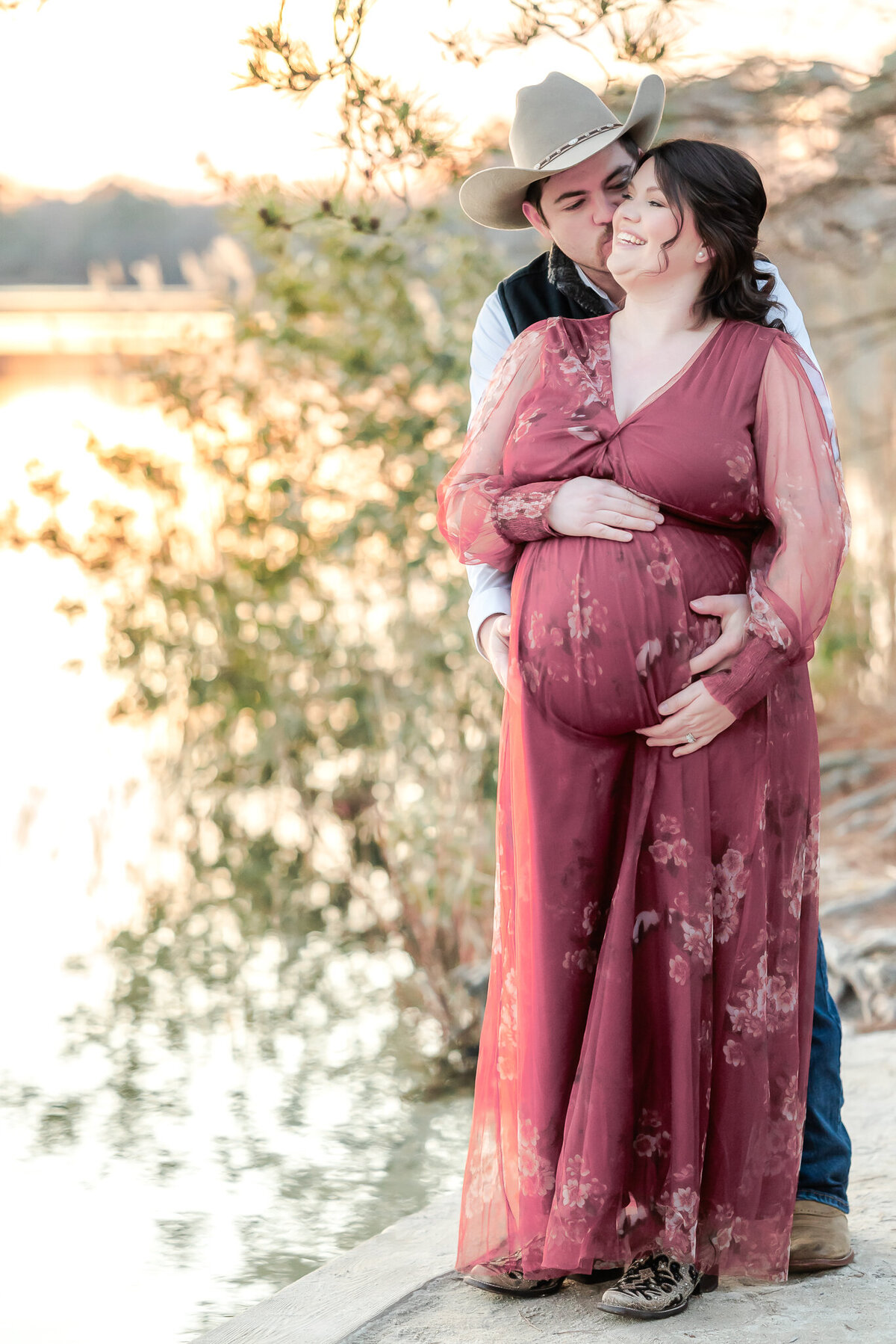 A man stands behind his pregnant wife and kisses her cheek during a maternity photography session in Chesapeake, VA. She wears a floor length, pink floral gown.