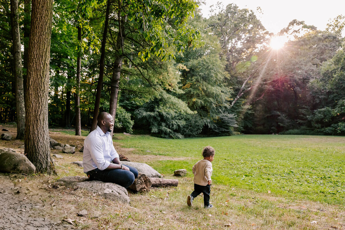 Black man in a pale blue dress shirt sits on a rock overlooking a flat meadow, with golden afternoon sun peaking through the leaves. His toddler runs into the grass just a few feet in front of him.