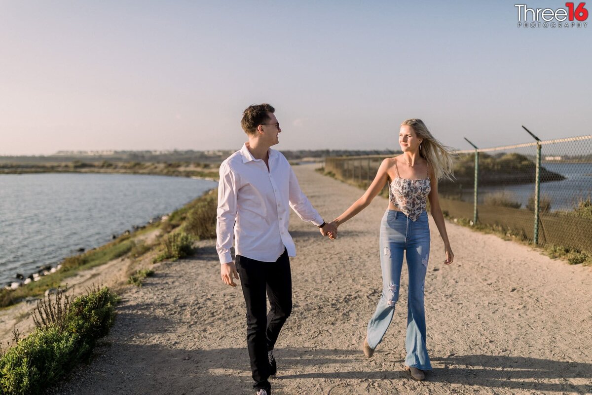 Engaged couple walk along a Bolsa Chica Ecological Reserve trail holding hands during photo shoot