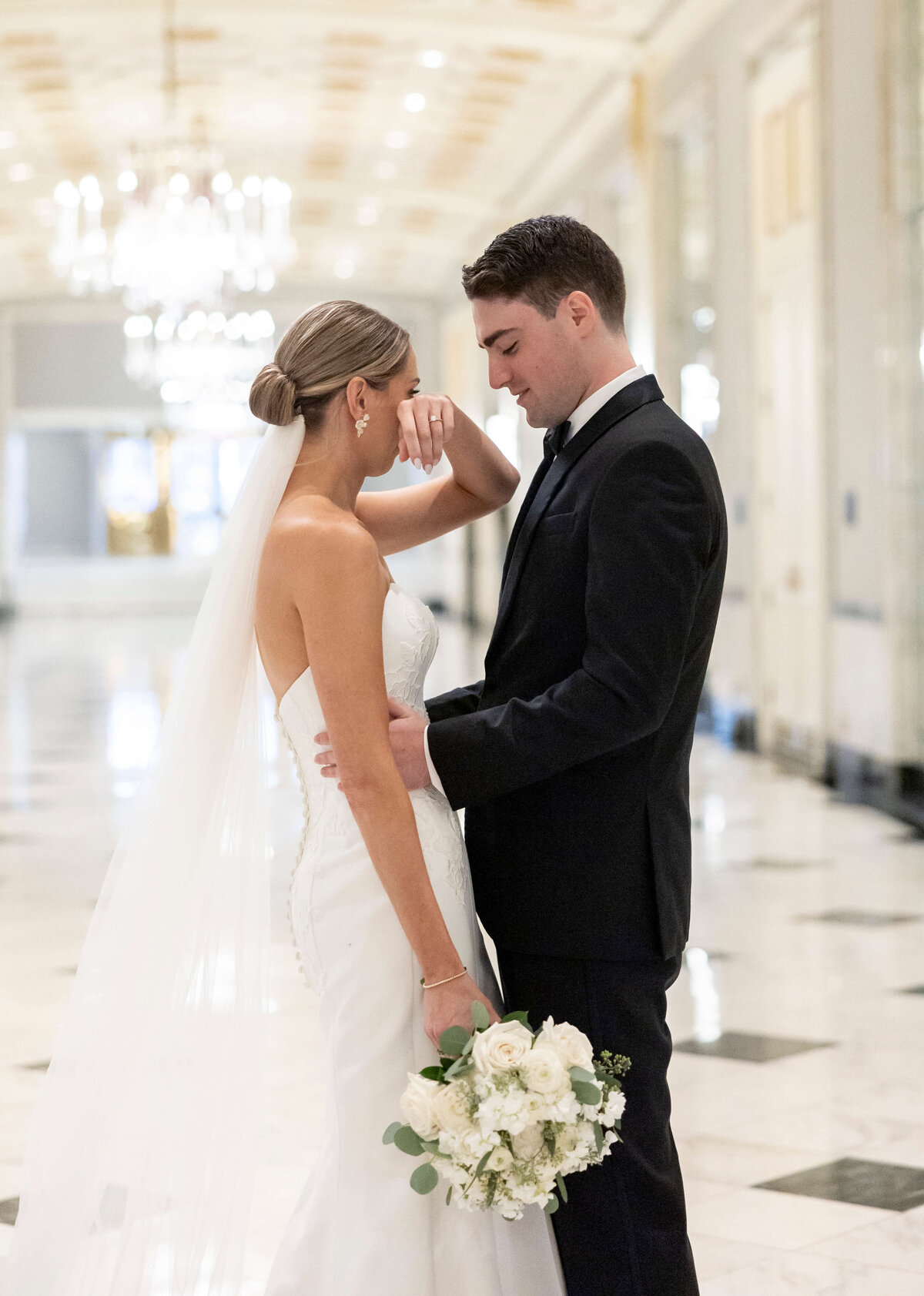 A bride and groom stand facing each other in an elegant hallway. The bride, in a strapless white gown and veil, holds a bouquet and wipes a tear. The groom is in a black suit. Chandeliers and ornate ceilings are visible in the background.