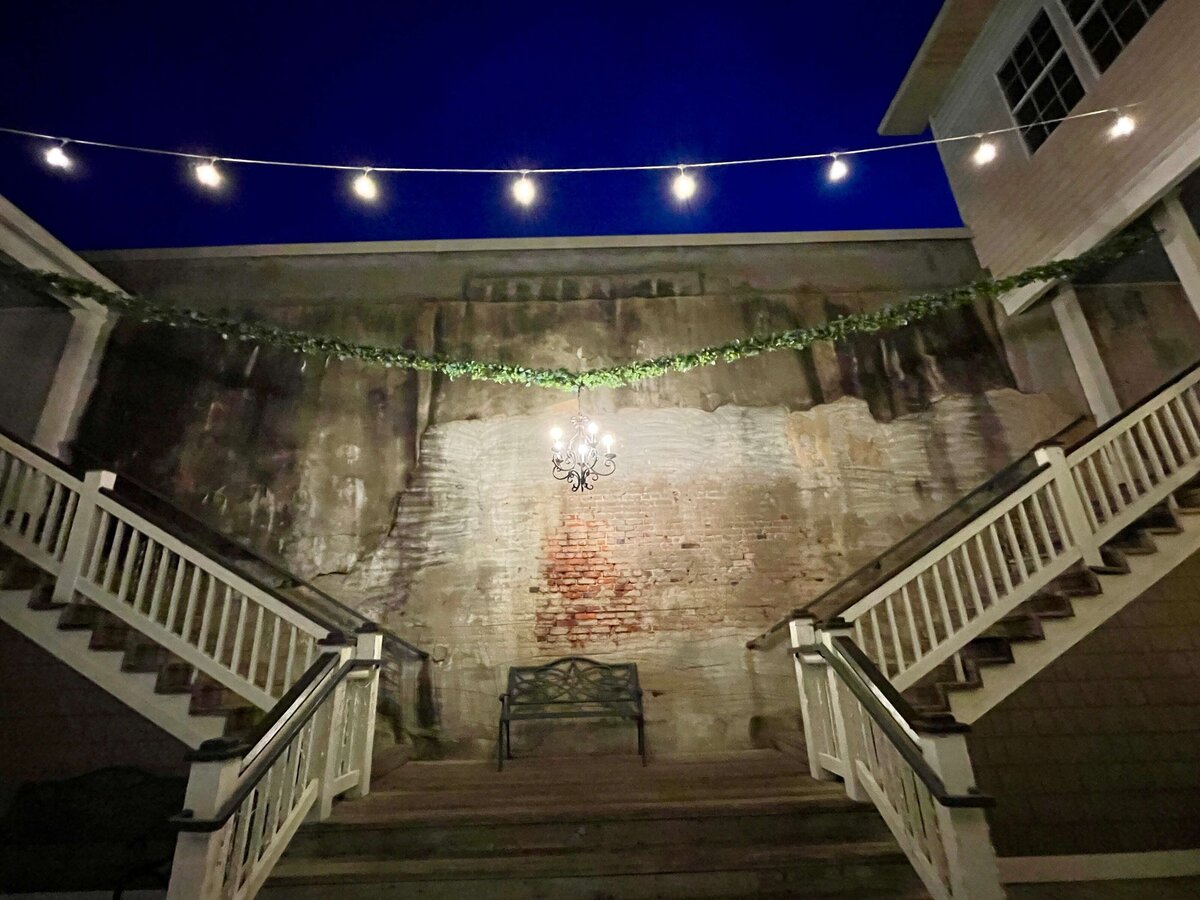 A nighttime scene of a stairway split in two, leading up to a brick wall with a bench beneath a hanging chandelier. String lights and greenery are draped above, with building exteriors on either side.
