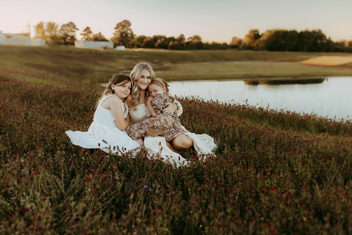 A woman and two young girls sit together in a field of wildflowers near a pond, with trees and buildings in the background at sunset—beautifully captured by a Warner Robins Family Photographer at Flat Creek.