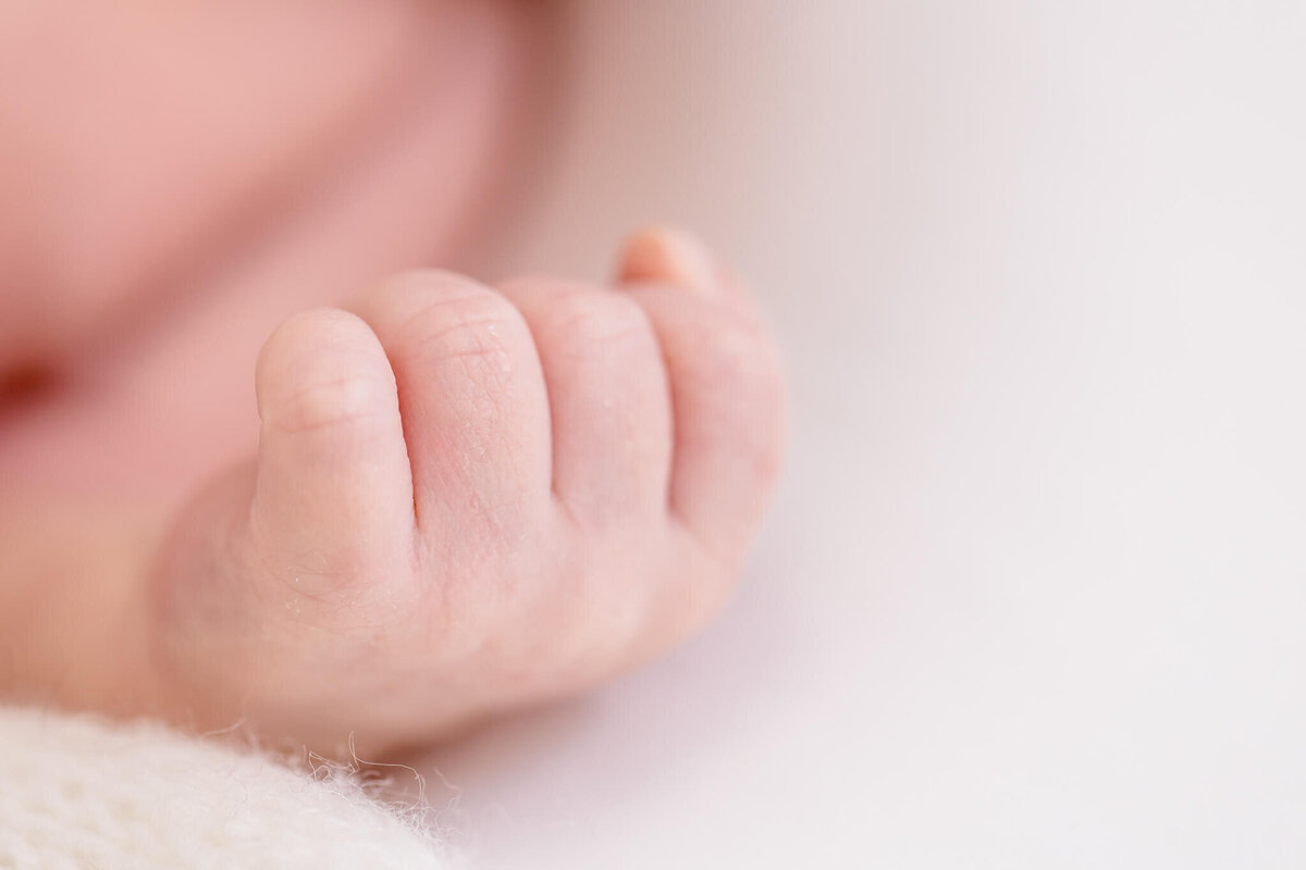 Close-up of a newborns hand with tiny fingers curled gently, resting on a soft white surface. The background is blurred, emphasizing the delicate features of the baby's skin.