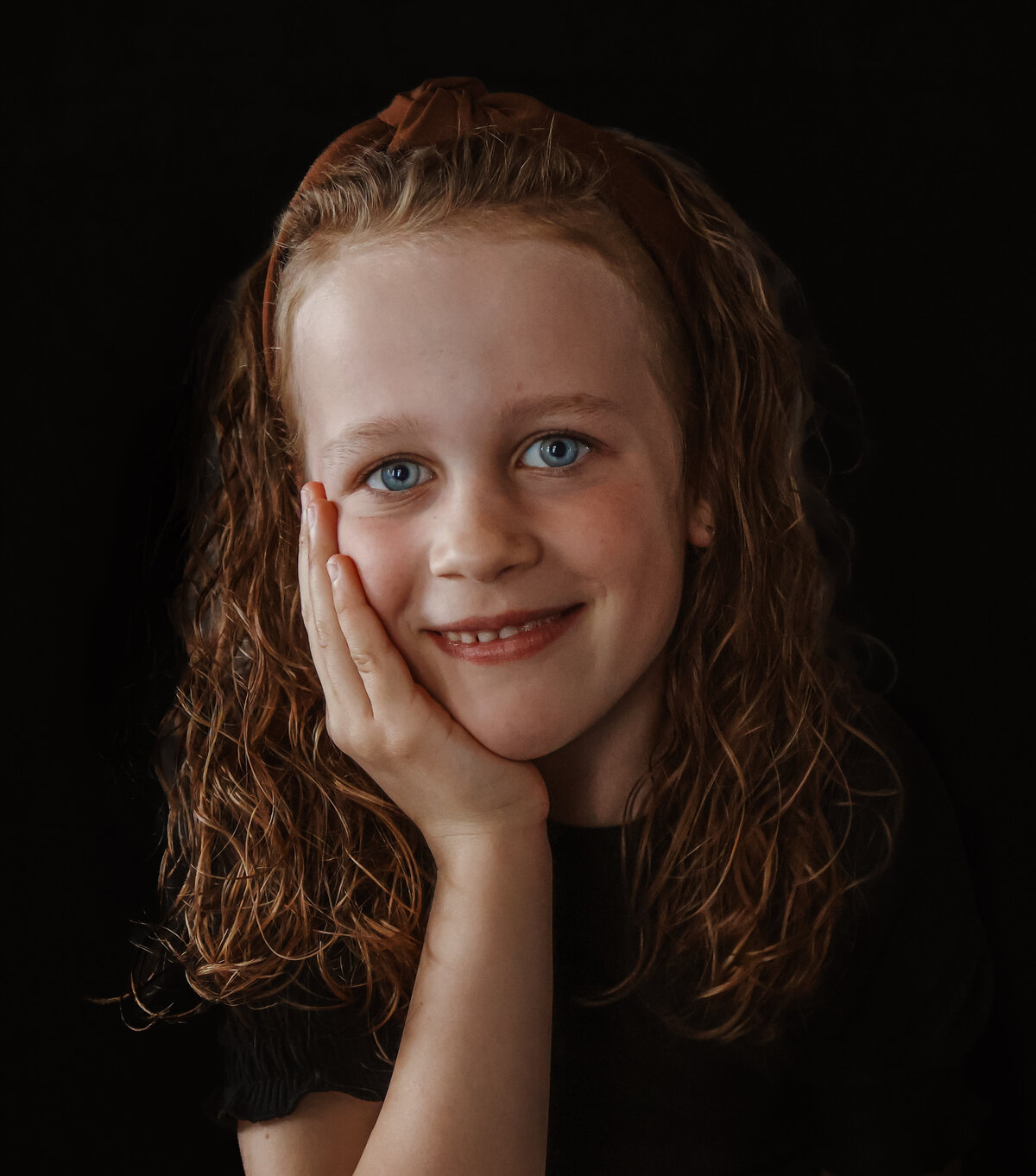 Een meisje met hand tegen haar wang en vrolijke glimlach, gefotografeerd tegen een donkere achtergrond
