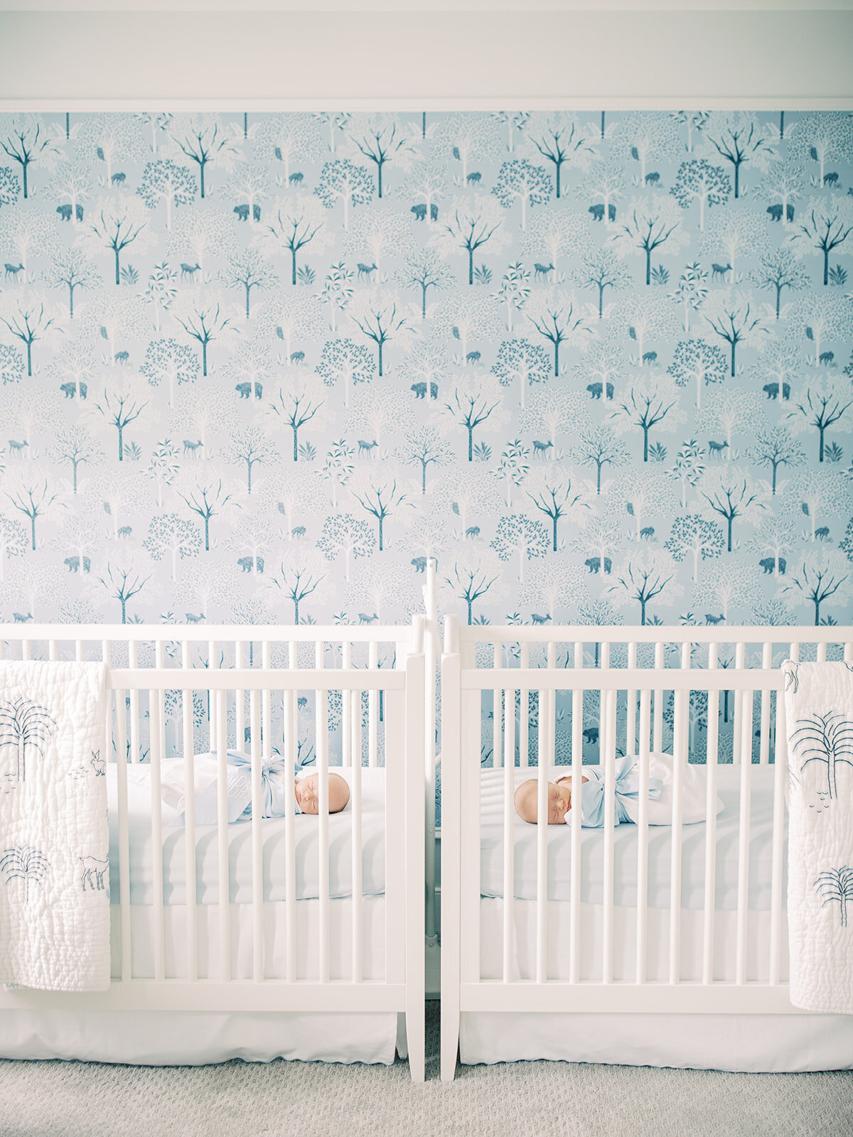 Twin baby boys sleep in their white cribs in their nursery with blue Serena & Lilly wallpaper.