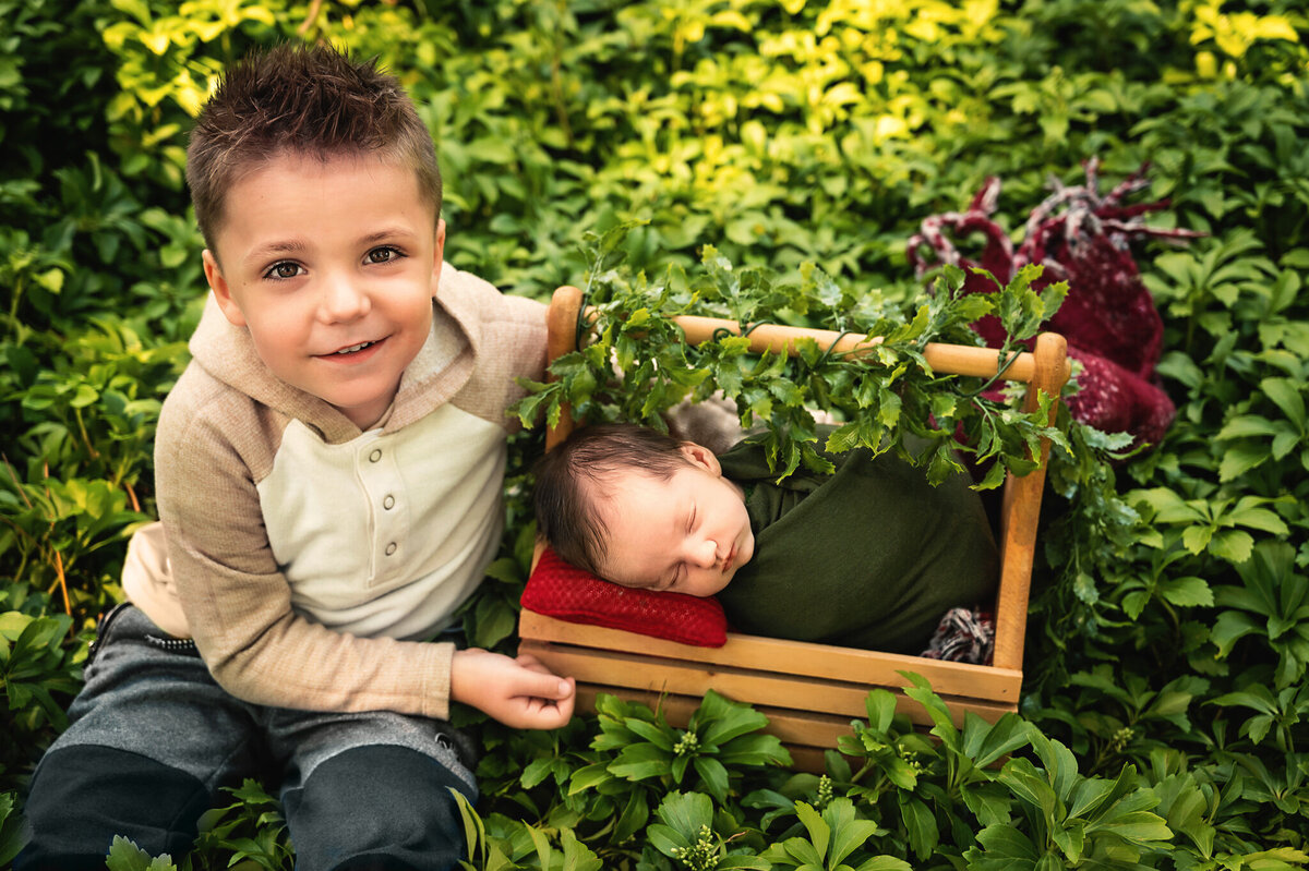 Brother smiling at camera sitting beside baby brother wrapped in a basket at outdoor photo session by Tamara Danielle Photography.