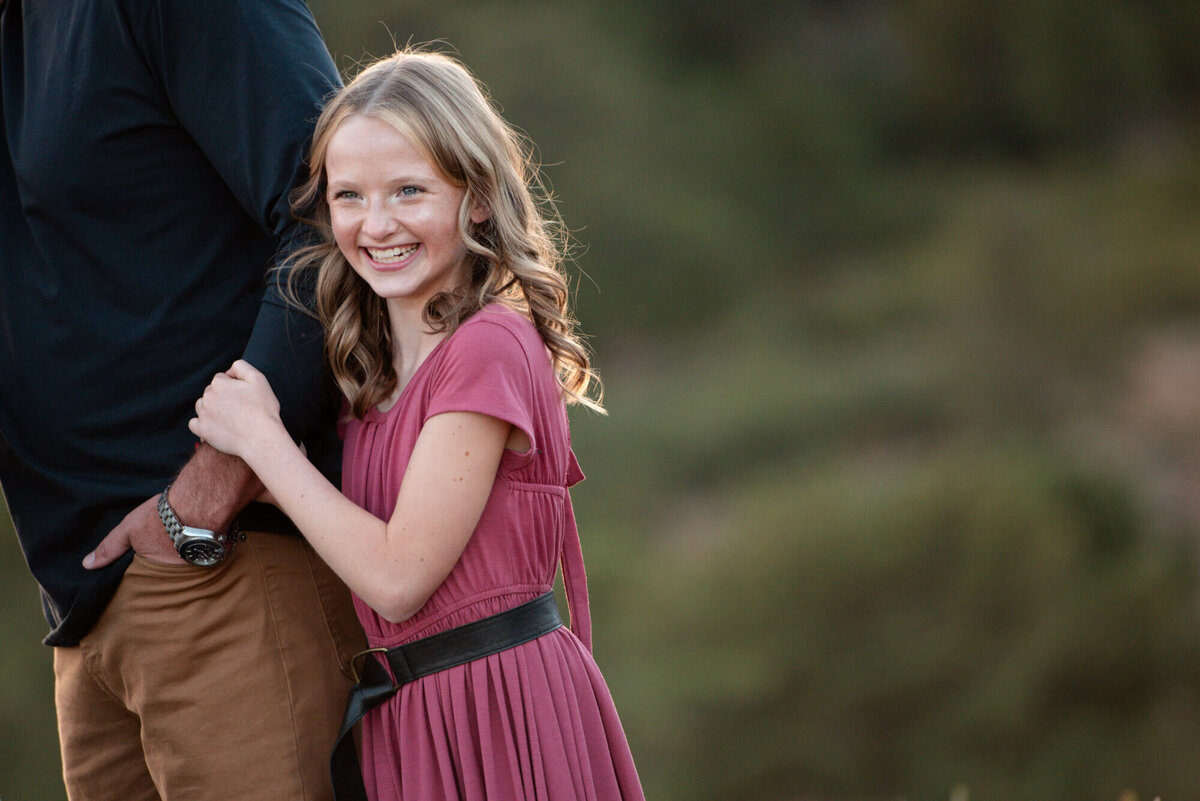 A young girl in a pink dress smiles big while hugging onto dad's arm