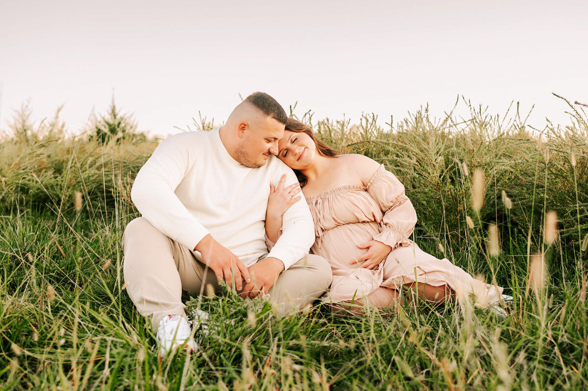 pregnant mom leaning on husbands shoulder sitting in grass at sunset during Branson MO maternity photography session