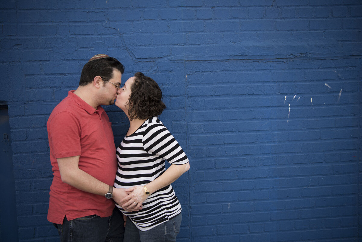 A pregnant couple kissing while they hold hands 