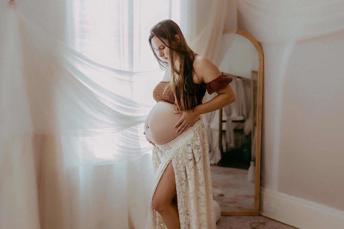 Pregnant woman in a lace skirt holds her belly while standing by a window with sheer curtains and a floor mirror in the background.