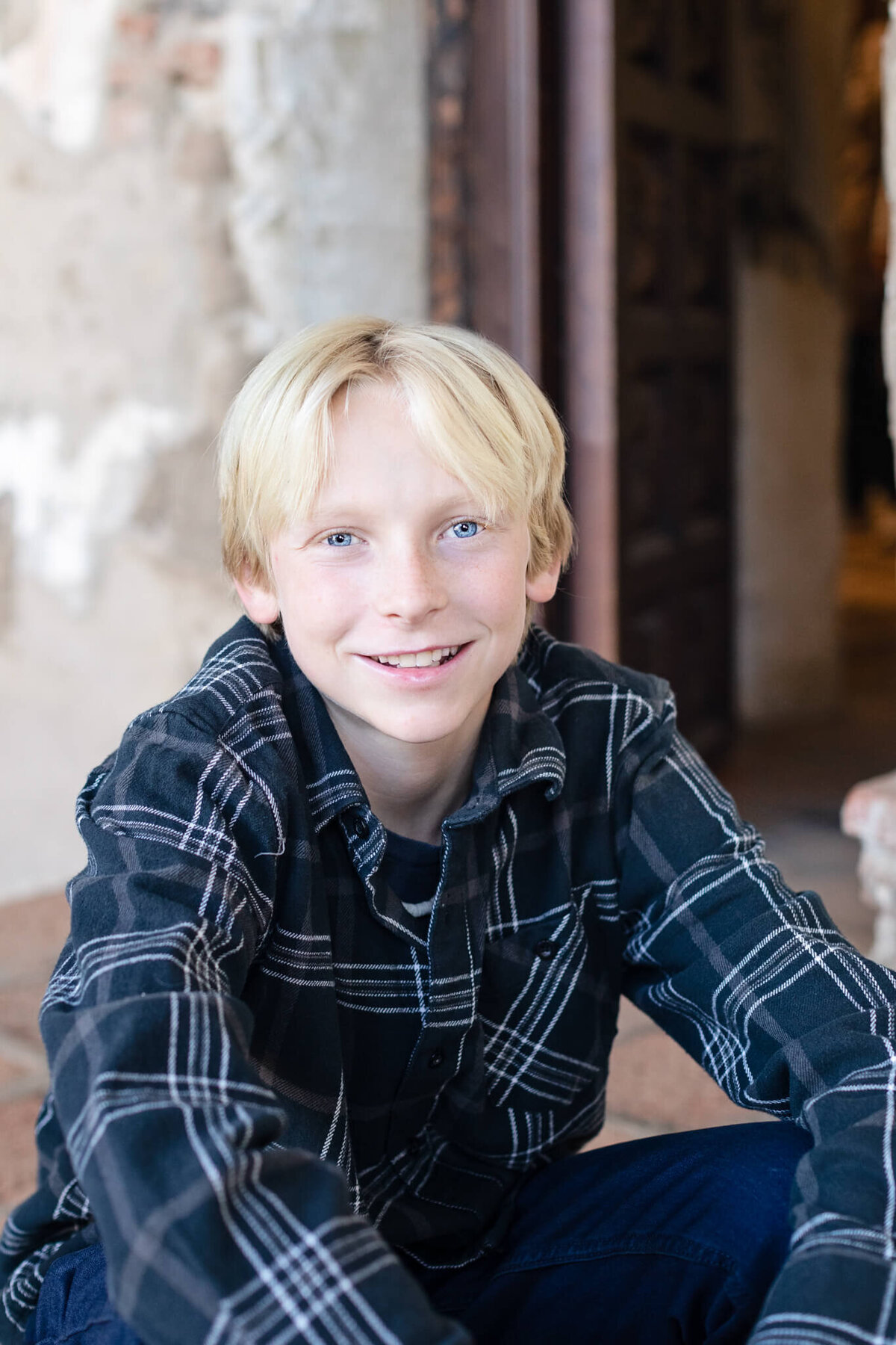 A close-up of a teenage boy with blonde hair and blue eyes wearing a dark blue flannel shirt and blue jeans looking at the camera at Mission San Juan Capistrano.