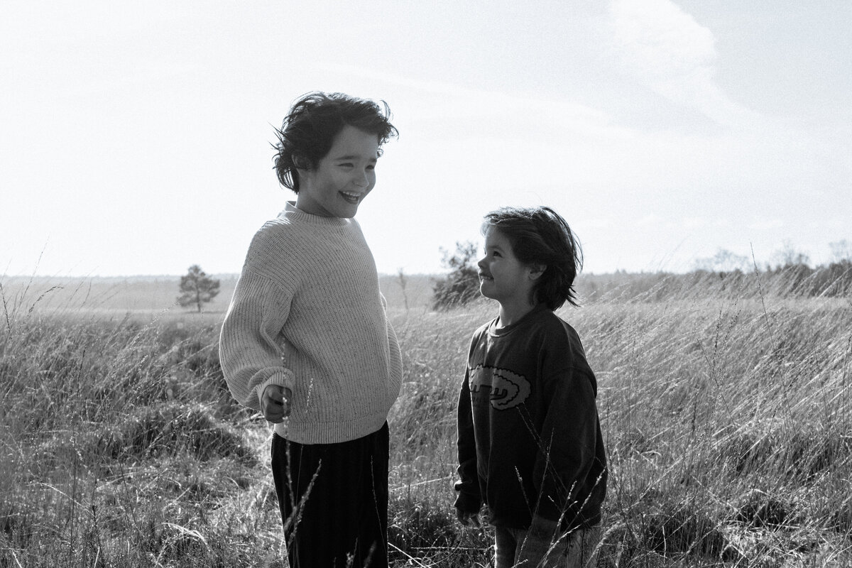 Twee kinderen staan in een veld, omringd door gras en bloemen, met een heldere lucht op de achtergrond.