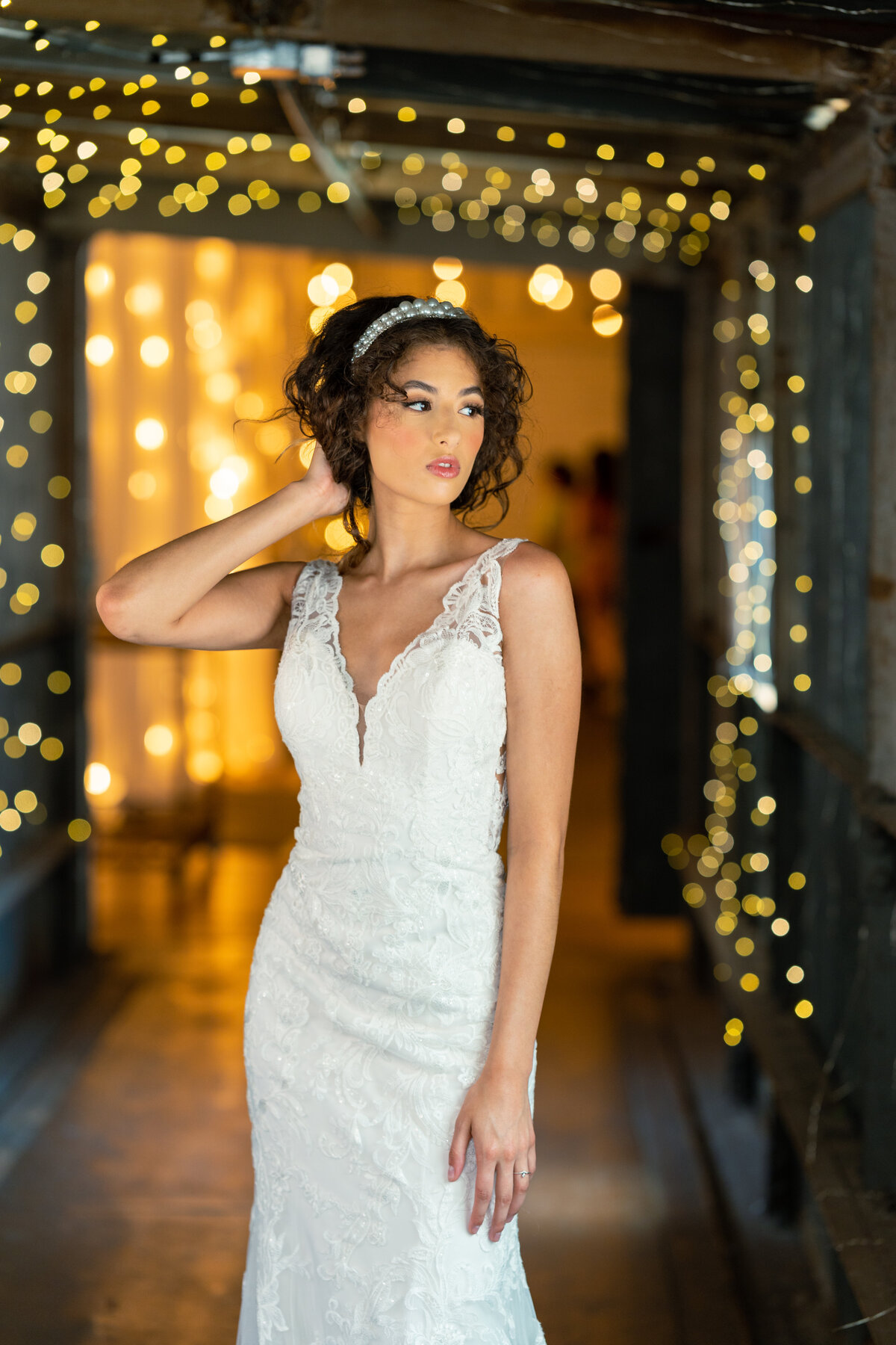 Curly-haired brunette model wearing lacey wedding dress with thick straps against a sparkly hall background.