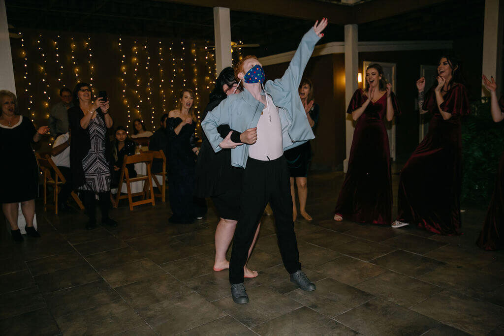 A person wearing a blue mask and light jacket strikes a playful pose on a dance floor while others around, dressed in formal attire, watch, clap, and laugh with string lights in the background.