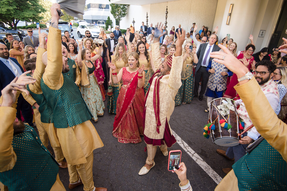 Energetic Indian wedding baraat celebration at Le Méridien Charlotte with groom in sherwani, bride in red lehenga, guests dancing, and dhol drummer. Captured by DeLong Photography, Charlotte Indian wedding photographers.