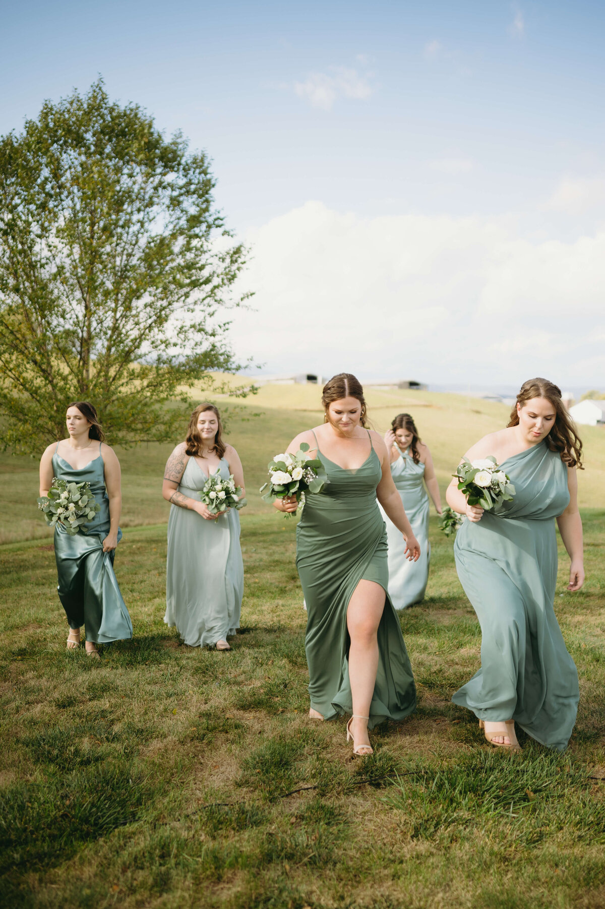 bridesmaids in green bridesmaids dresses walking through a field in Sunny Slope Farms for a summer wedding
