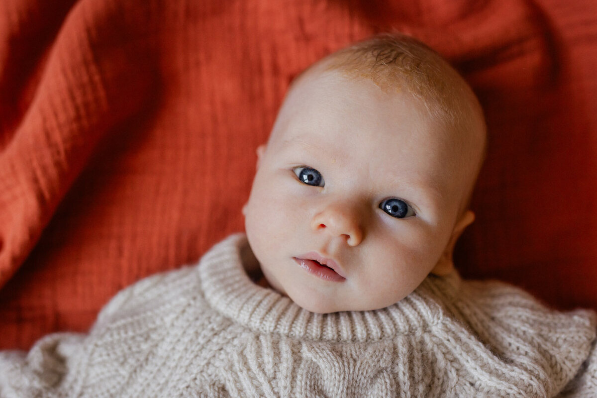 newborn with blue eyes and dressed in cable-knit sweater laying on orange swaddle blanket at home and looking at camera