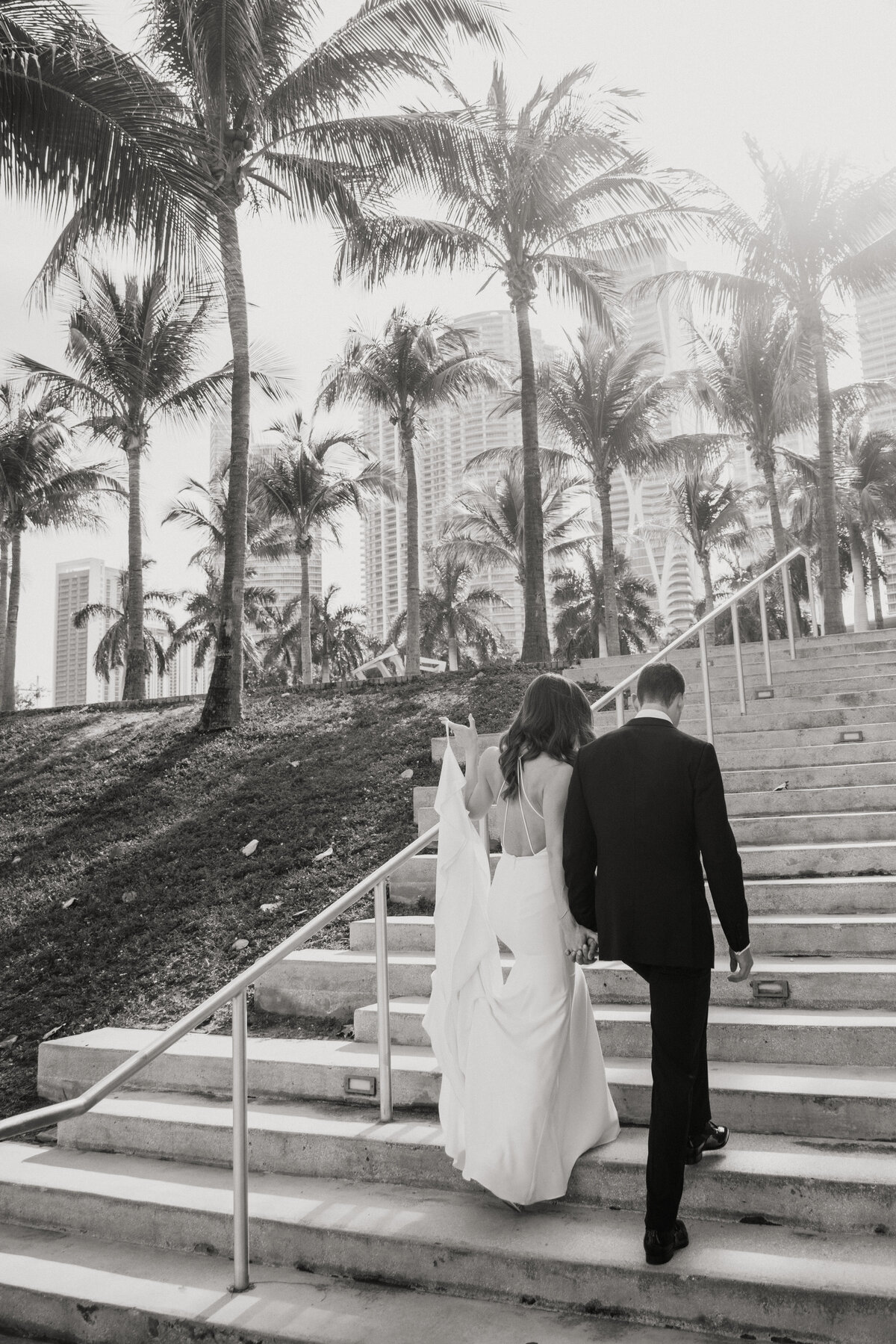 Bride and groom walking up stairs with palm trees and Miami skyline in the background.