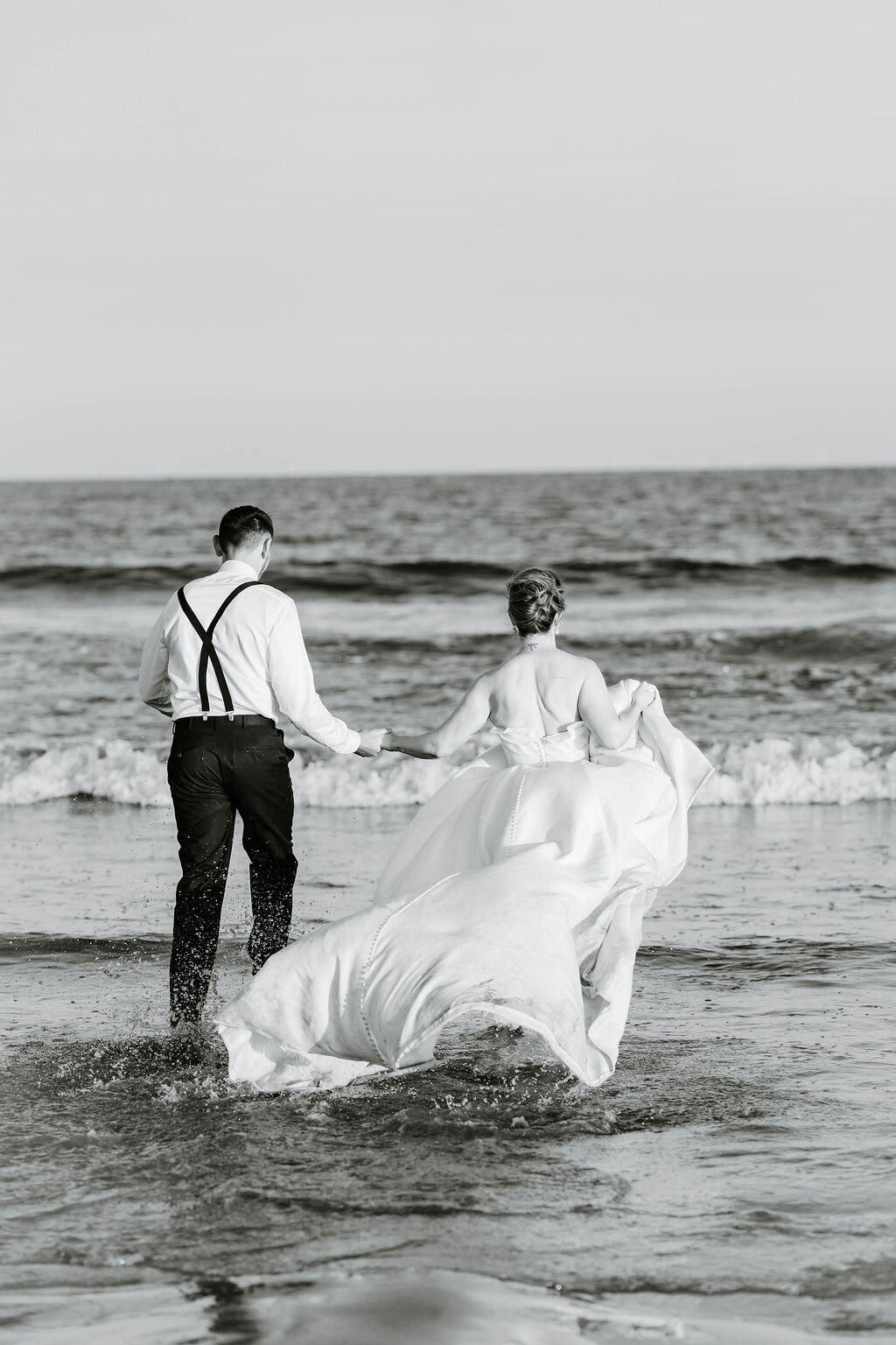 Rhode Island Elopement | A couple, dressed in wedding attire, walks hand in hand toward the ocean. The bride's gown trails in the water.
