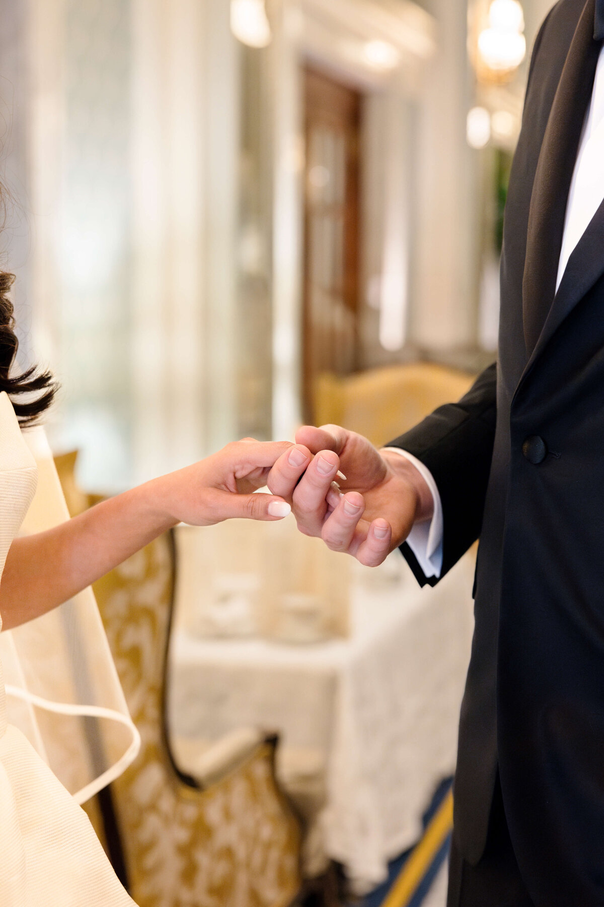 A couple dressed in formal attire holds hands. The woman is in a white dress, and the man is in a black suit. They are indoors, with a blurred background that includes a decorated table and chairs.