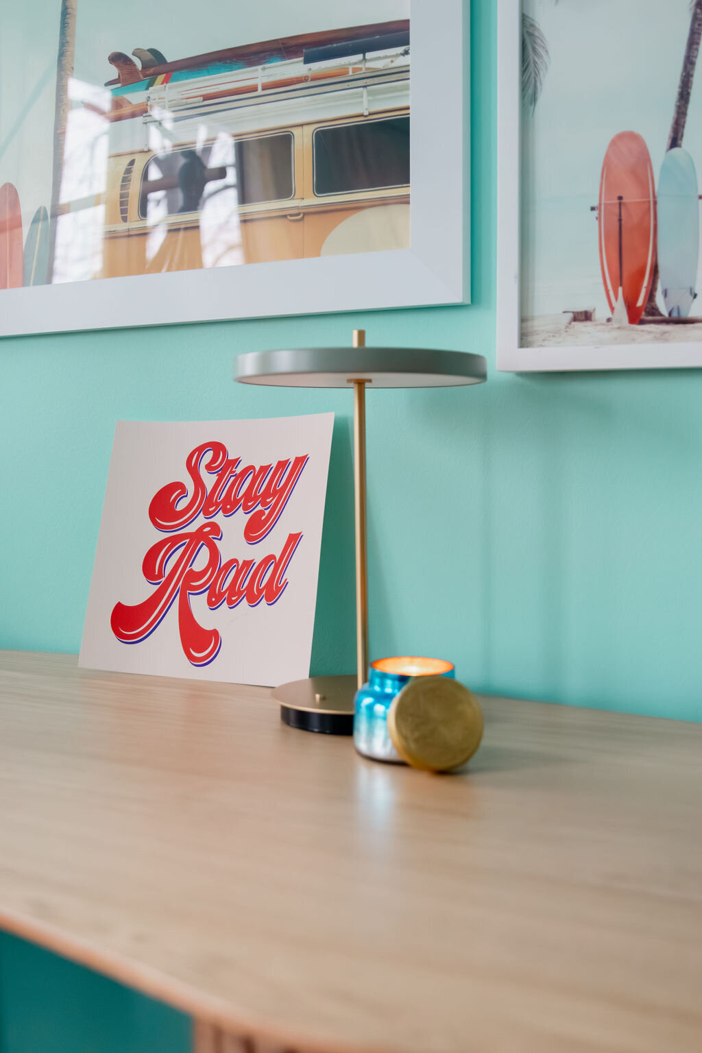 A close-up of a light-colored Umage Audacious desk featuring a gray Umage Asteria Move Portable Table Lamp with a gold stem and a circular shade. Next to the lamp, a postcard with the red script text "Stay Rad" leans against the wall. A small, round candle with a patterned ceramic base and a gold lid sits near the lamp's base. Above, two framed beach-themed prints hang on the light turquoise wall. One depicts a vintage van with surfboards, and the other shows surfboards standing on a beach. The lighting is soft and focused on the objects on the desk.