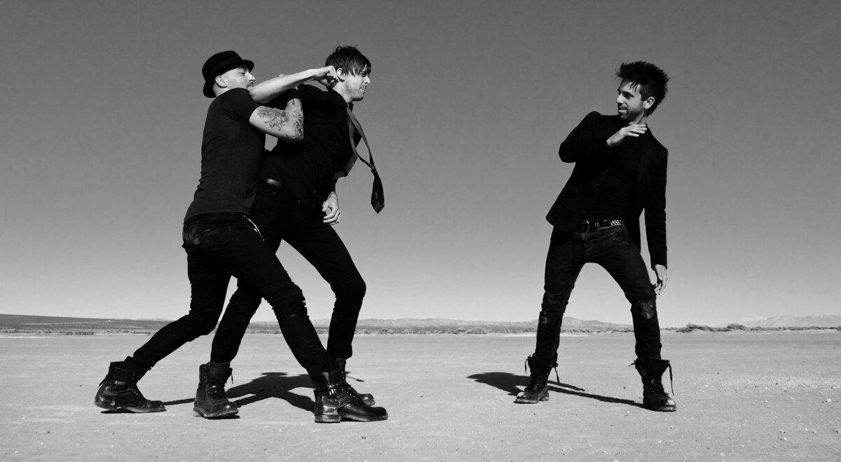 Black and white band portrait Angels Landing trio dressed in black one member holding another back as he lunges toward lead singer El Mirage