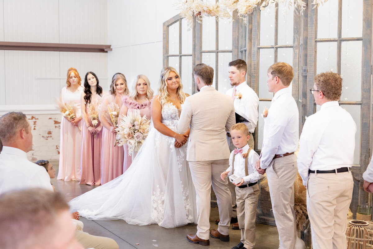 The bride and groom stand together at the altar, gazing into each other’s eyes as the wedding officiant begins the ceremony. Bridesmaids in elegant blush dresses and groomsmen in coordinating attire create a picture-perfect backdrop.